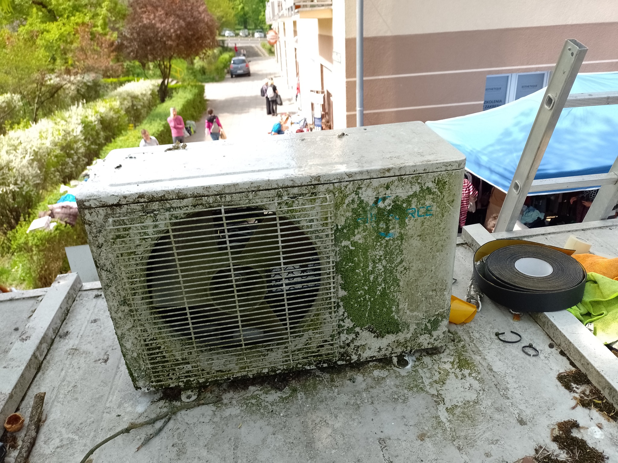 Zewnętrzna jednostka klimatyzacji pokryta mchem i zabrudzeniami, stojąca na dachu budynku, obok drabina i akcesoria serwisowe, widok z góry na otoczenie.