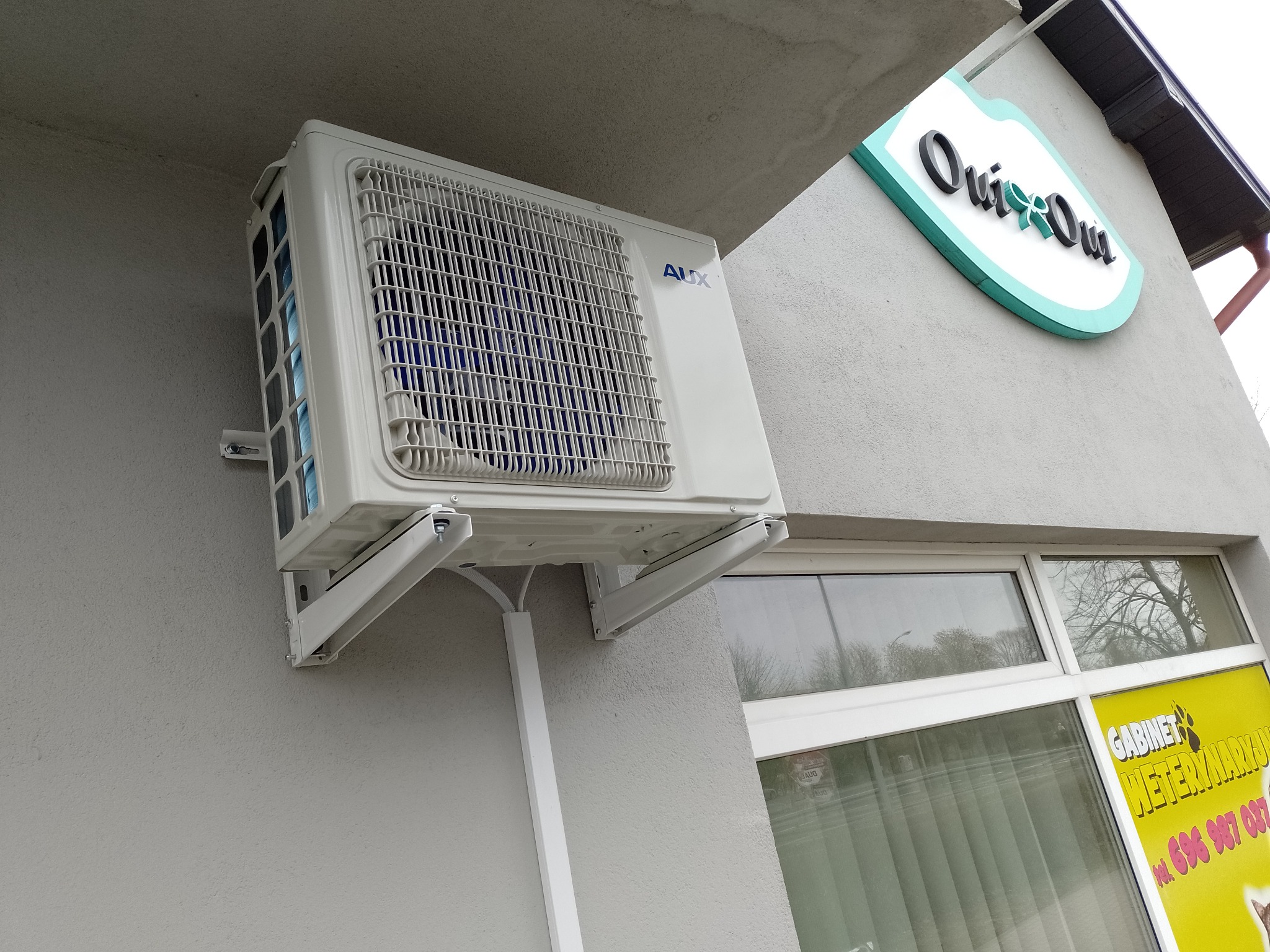 Zewnętrzna jednostka klimatyzacji AUX zamontowana na elewacji budynku, widoczna instalacja elektryczna w białej listwie, okno z reklamą gabinetu weterynaryjnego w tle.