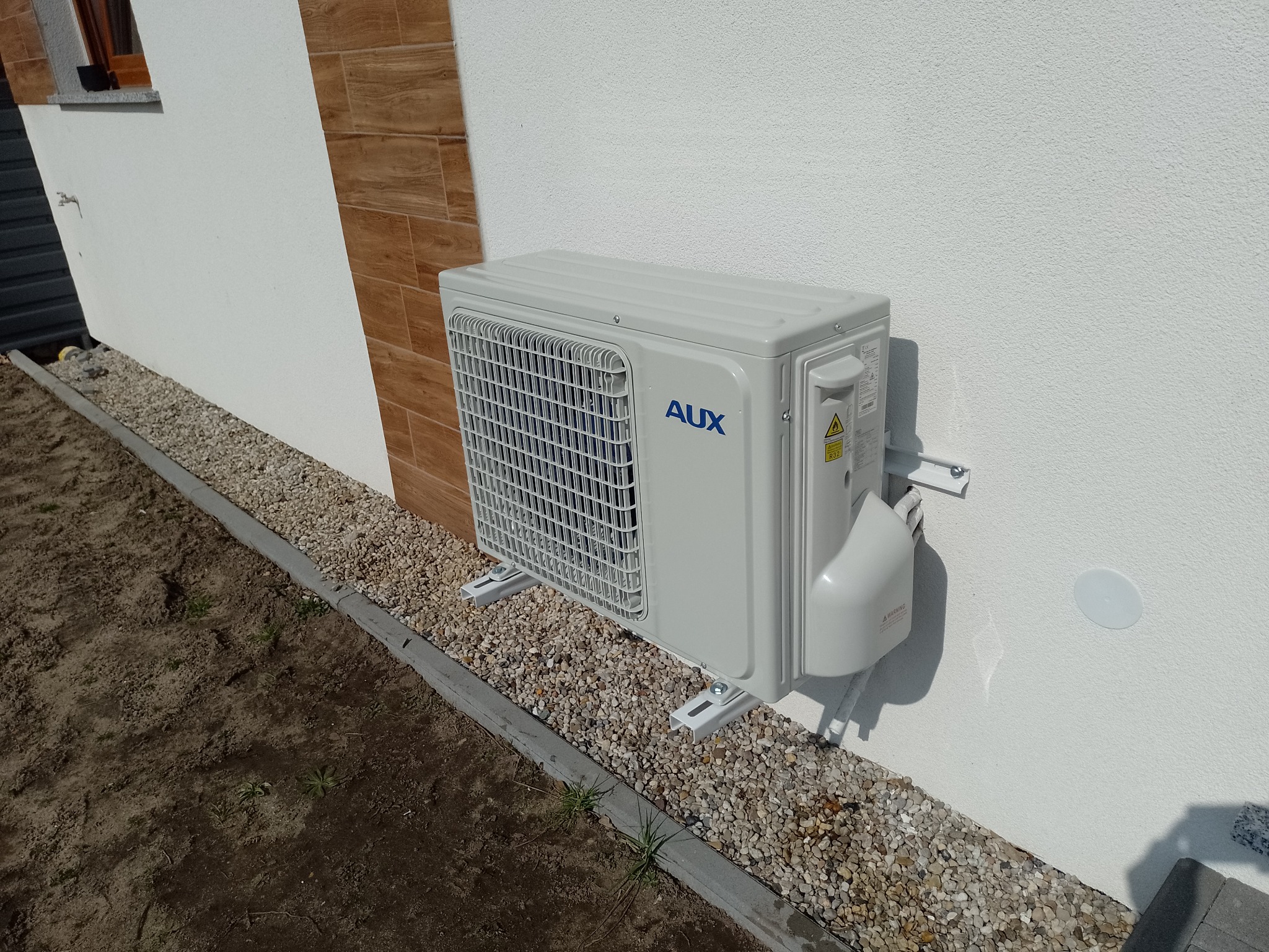 Zewnętrzna jednostka klimatyzacji AUX zamontowana na białej ścianie domu, widoczne kamienie ozdobne wokół podstawy urządzenia.