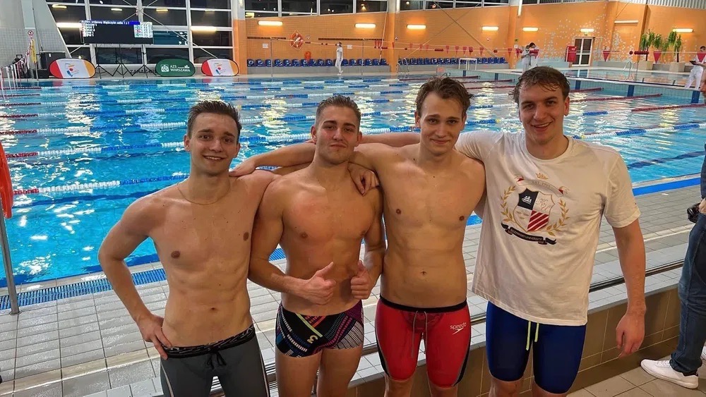 Grupa czterech młodych mężczyzn w strojach kąpielowych i koszulce, pozujących na tle basenu z niebieską wodą i torami pływackimi.