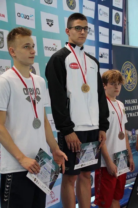 Trzech młodych pływaków z medalami na podium w Warszawie, trzymających dyplomy, z logotypami sponsorów w tle.
