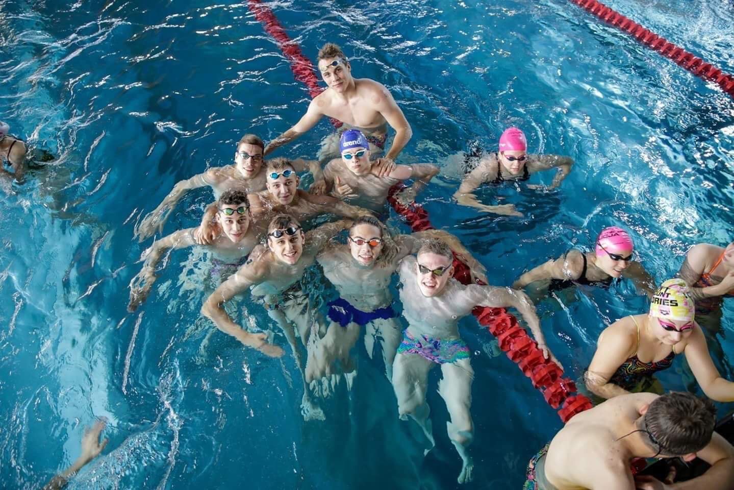 Grupa młodych pływaków w okularach i czepkach, uśmiechniętych, unoszących się na wodzie w basenie z czerwonymi torami.