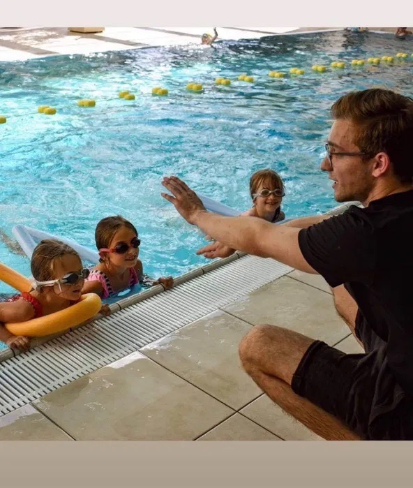 Instruktor pływania w okularach, kuca przy basenie, pokazując gesty trójce dzieci w okularach pływackich, z piankowymi makaronami i deskami, unoszącymi się na wodzie z żółtymi bojami.
