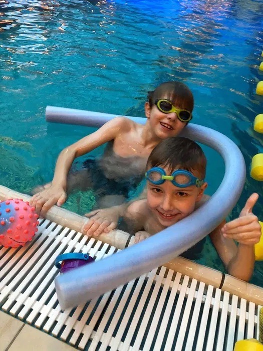 Dwóch uśmiechniętych chłopców w okularach do pływania, z makaronem do pływania w basenie, jeden pokazuje kciuk w górę.