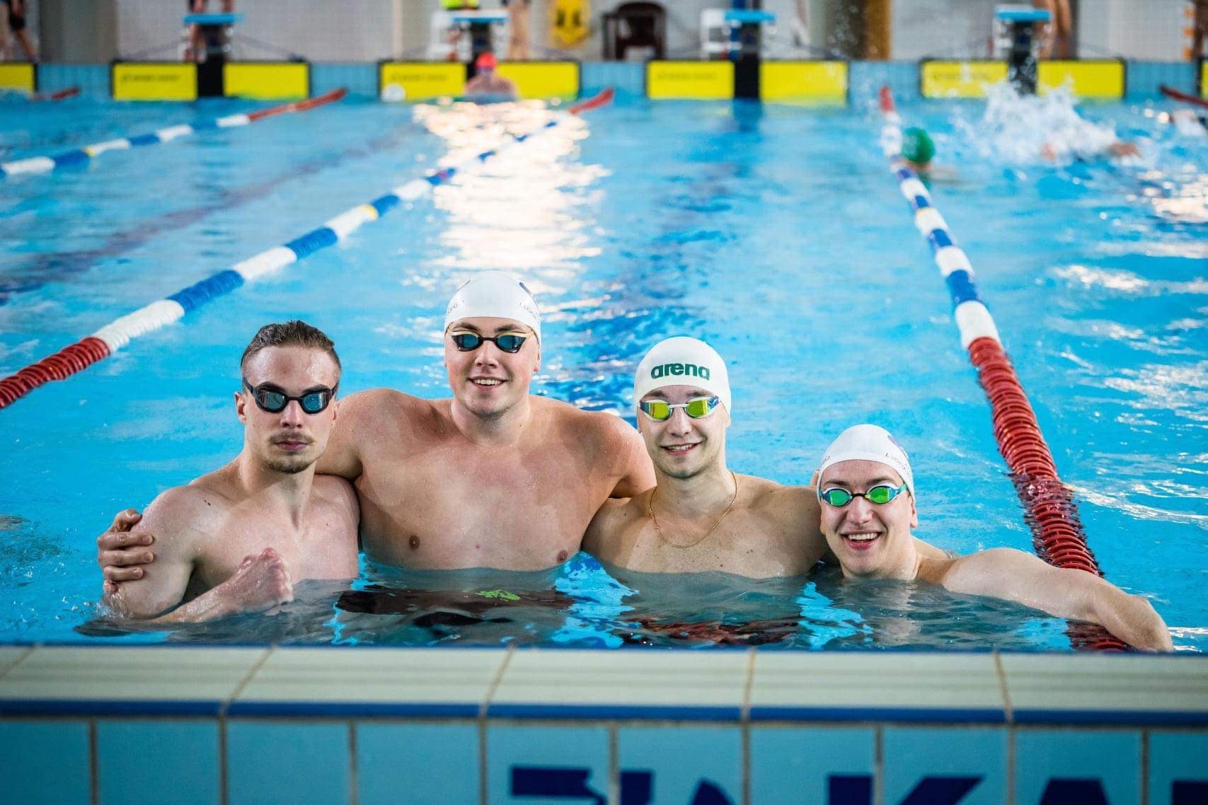 Grupa czterech młodych pływaków w czepkach i okularach, stojących w basenie, uśmiechniętych, z widocznymi linkami torów i osobami pływającymi w tle.