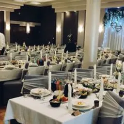 Sala bankietowa przygotowana na uroczystość, stoły z białymi obrusami i szarymi pokrowcami na krzesłach, dekoracje kwiatowe i serca na oknie, kelner w tle.