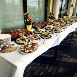 Stół cateringowy z białym obrusem, zastawiony różnorodnymi przekąskami, mini-burgerami, koreczkami i dekoracjami kwiatowymi w żółtych doniczkach, ustawiony na zewnątrz przy kolumnach.