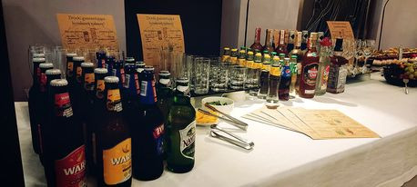 Stół z napojami alkoholowymi i bezalkoholowymi, szklankami, szczypcami do lodu i menu na białym obrusie, przygotowany na przyjęcie.