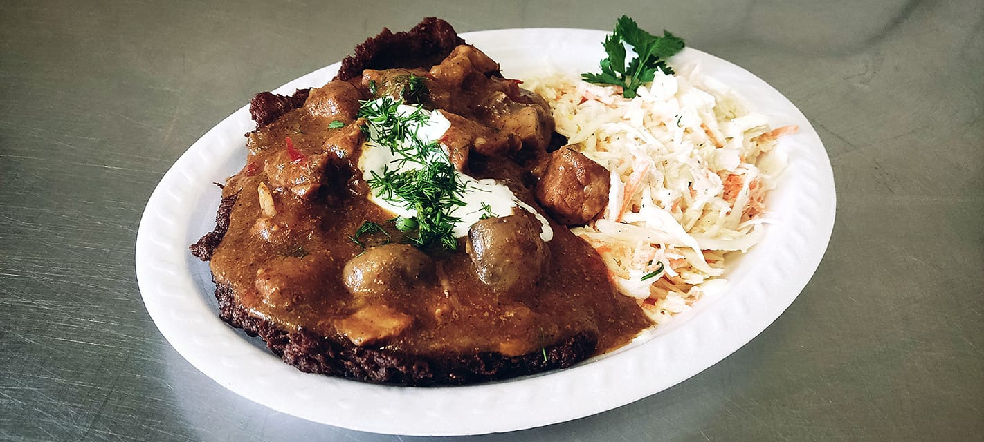 Placek ziemniaczany po węgiersku z gulaszem, kleksem śmietany, koperkiem i surówką z białej kapusty na białym talerzu.