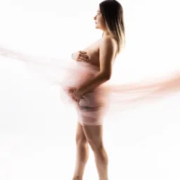 Kobieta w ciąży, profil, owinięta w delikatny, różowy materiał, na białym tle, z widocznym tatuażem palmy na stopie, delikatne oświetlenie.