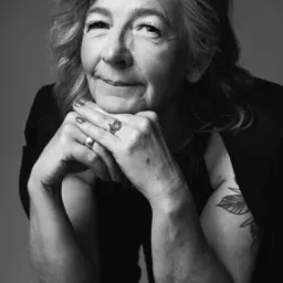 Portret studyjny kobiety w średnim wieku w czerni i bieli, z rękami podpartymi pod brodą, pierścionkami i tatuażem na ramieniu.