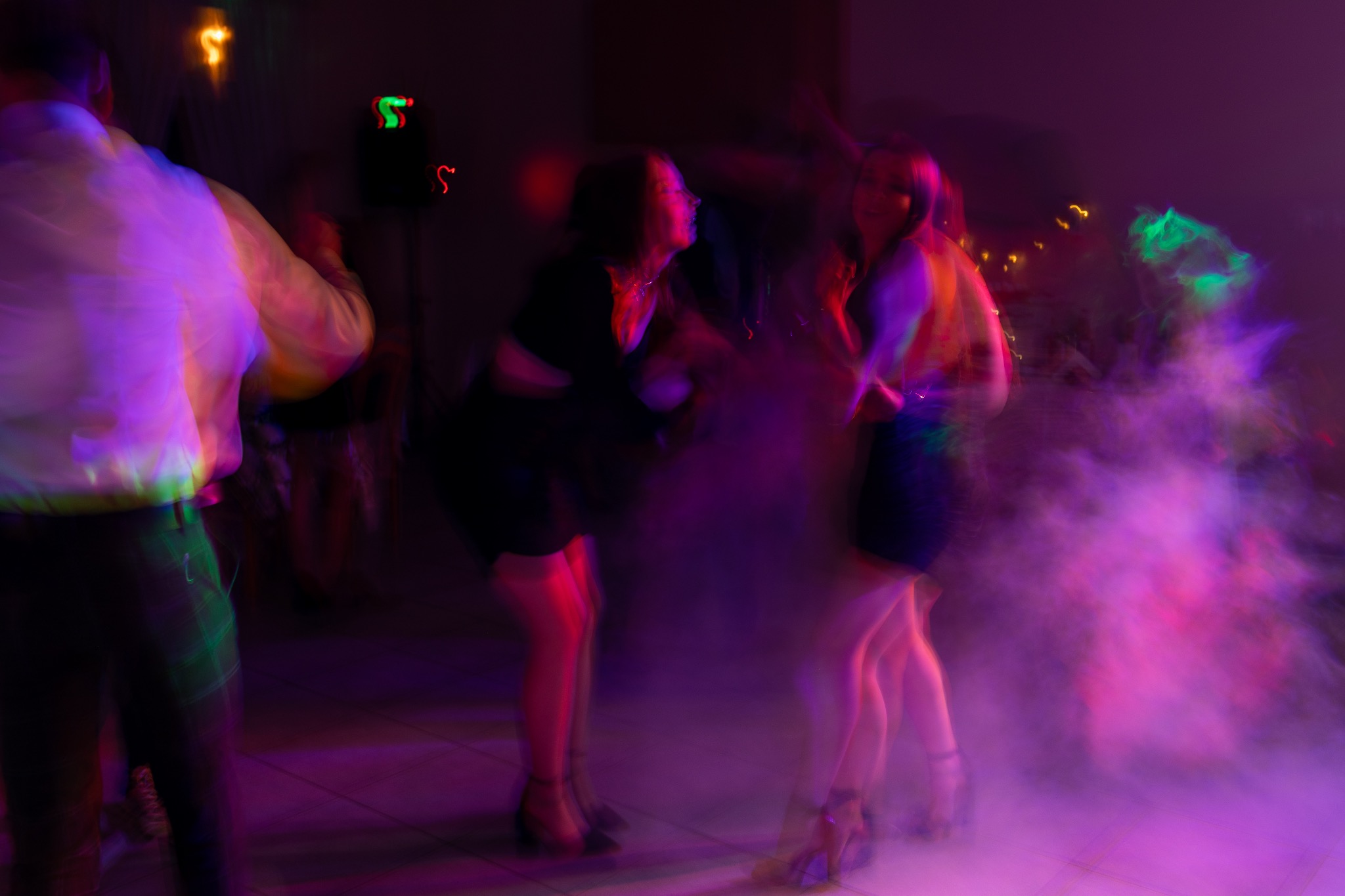 Rozmazane ujęcie tańczących kobiet na parkiecie w fioletowym oświetleniu, z elementami dekoracyjnymi na ścianie i efektem dymu.
