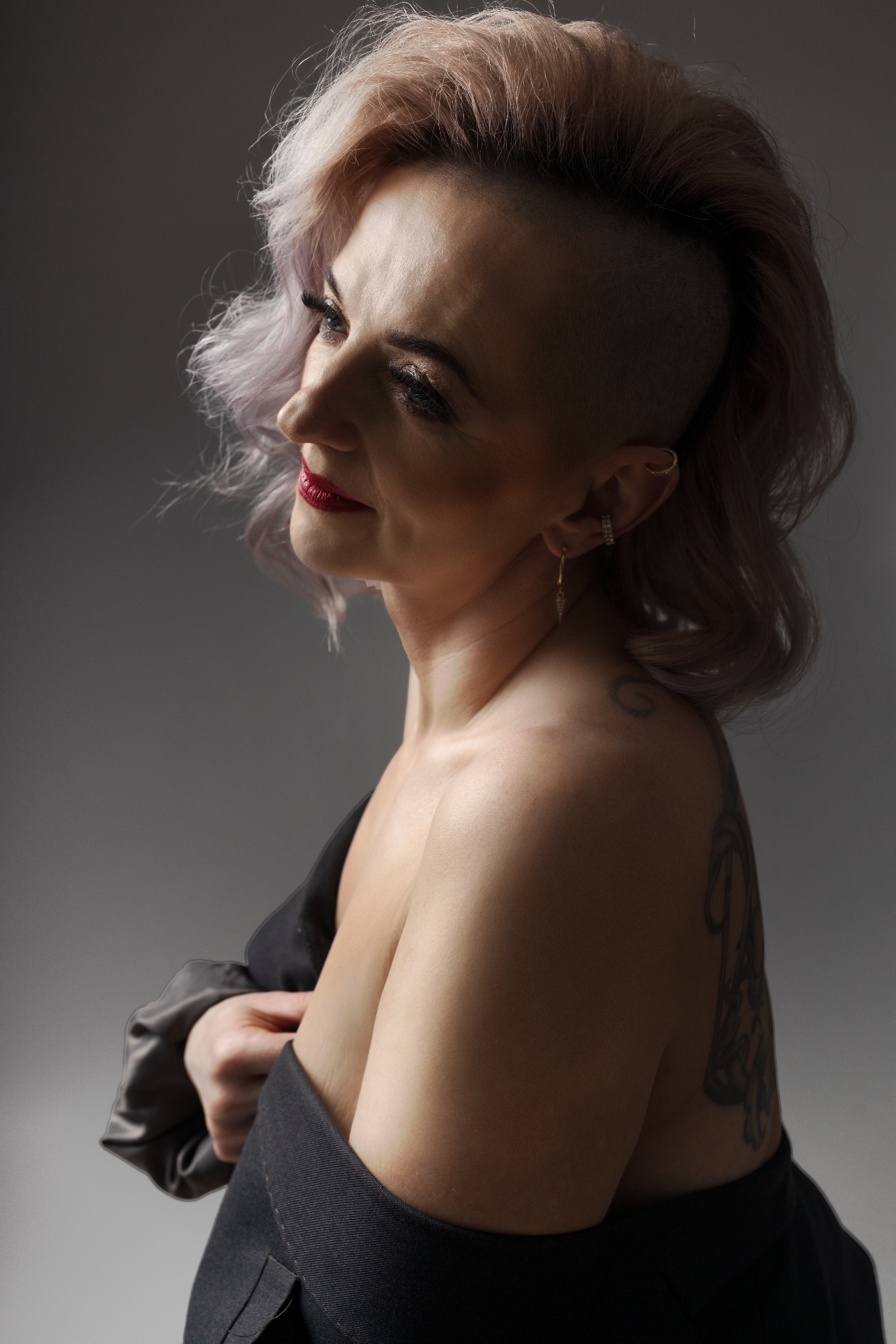 Portret kobiety z asymetryczną fryzurą, różowymi włosami i tatuażem na ramieniu, ubrana w czarną marynarkę, z delikatnym makijażem i kolczykami, spoglądająca w bok na szarym tle.