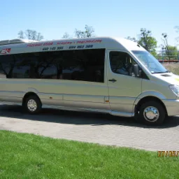 Jasny bus marki Mercedes Sprinter z przyciemnianymi szybami i napisem 'PRZEWÓZ OSÓB - krajowy i zagraniczny' oraz numerami telefonów, zaparkowany na kostce brukowej obok trawnika w słoneczny dzień.