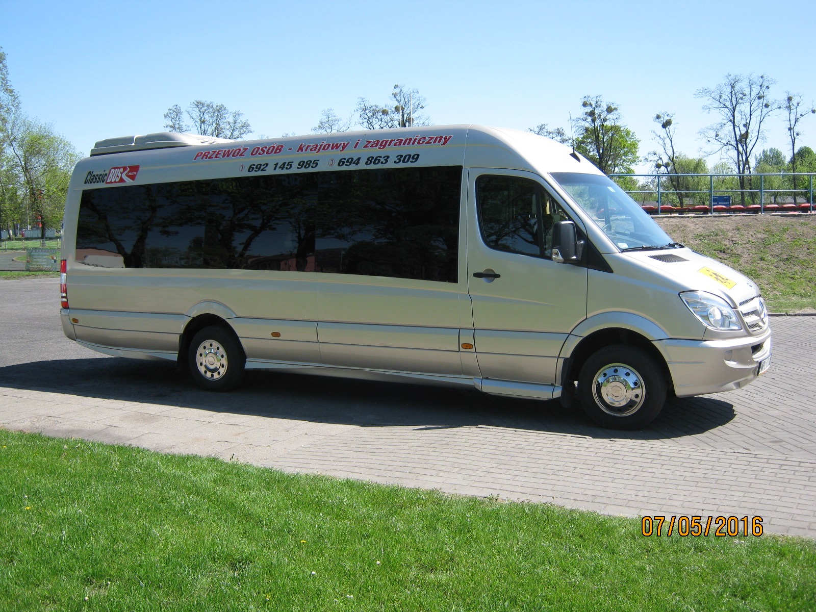 Jasny bus marki Mercedes Sprinter z przyciemnianymi szybami i napisem 'PRZEWÓZ OSÓB - krajowy i zagraniczny' oraz numerami telefonów, zaparkowany na kostce brukowej obok trawnika w słoneczny dzień.