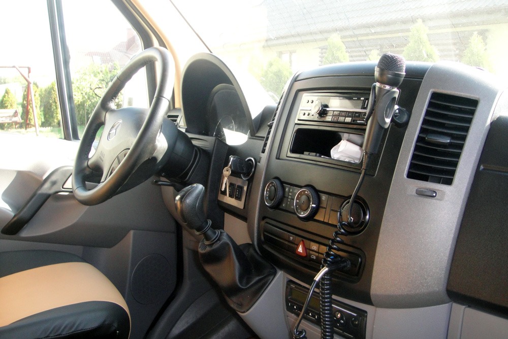 Wnętrze samochodu dostawczego Mercedes-Benz z mikrofonem zamocowanym na desce rozdzielczej, widoczna kierownica, skrzynia biegów i panel sterowania.