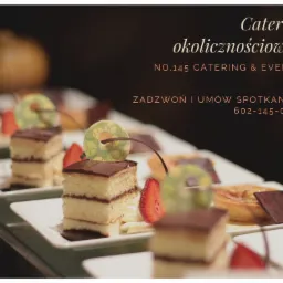 Ustawione w rzędzie porcje ciasta z czekoladową polewą, truskawkami i dekoracyjnymi krążkami na białych talerzykach, w tle dynia i tekst reklamowy firmy cateringowej.