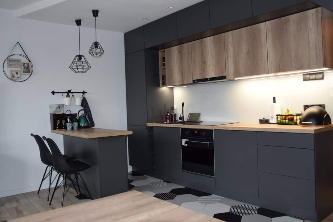 Nowoczesna kuchnia z szarymi szafkami dolnymi i drewnopodobnymi górnymi, oświetleniem LED pod szafkami, blatem z jasnego drewna i heksagonalną podłogą w odcieniach szarości i czerni.