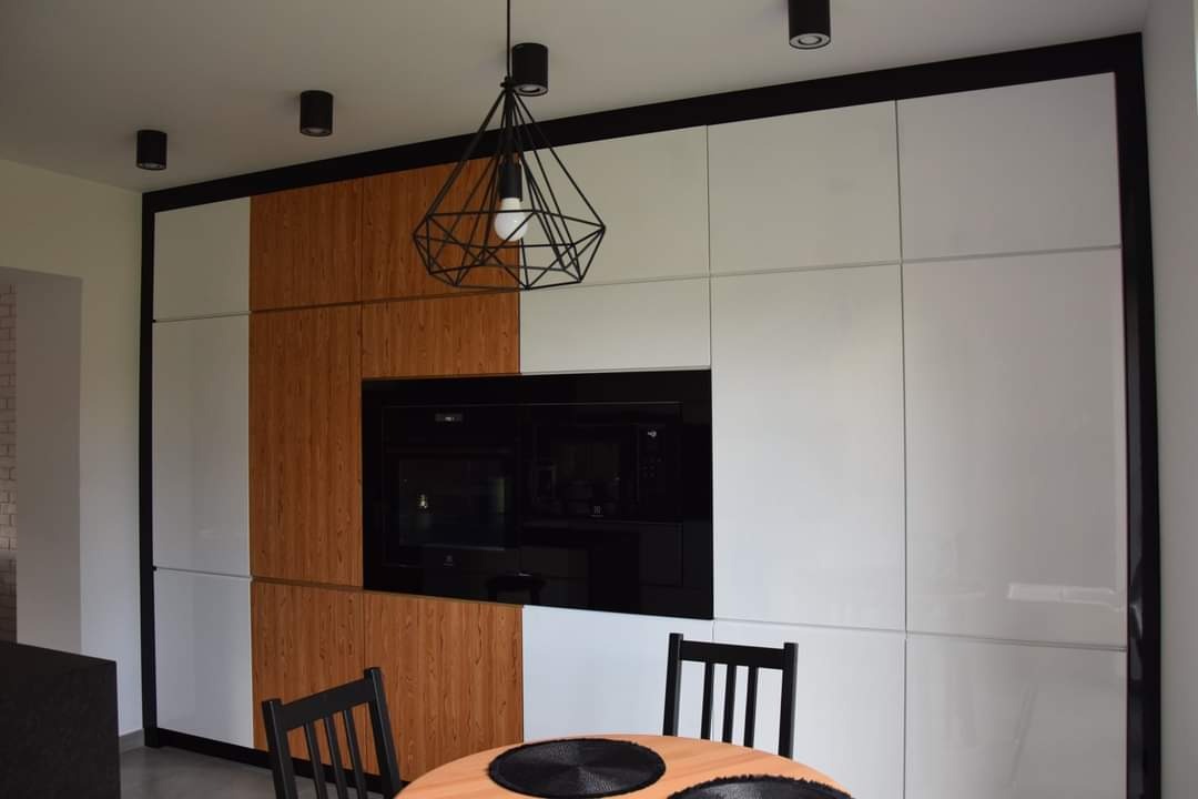 Nowoczesna kuchnia z białymi i drewnopodobnymi frontami, zabudowana piekarnikiem i wiszącą geometryczną lampą nad okrągłym stołem z czarnymi podkładkami.