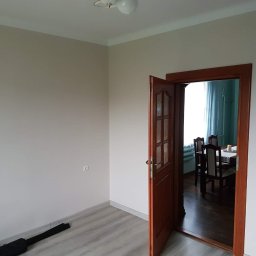 Paweł Stanek - Jasne wnętrze po remoncie: ściany pomalowane na beżowo, drewniane drzwi uchylone, widoczny fragment podłogi z paneli i wnętrze drugiego pokoju z zasłonami i stołem z krzesłami.