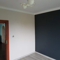 Paweł Stanek - Pomieszczenie po remoncie: ściany pomalowane na biało i granatowo, drewniane drzwi z szybkami, szare panele podłogowe, sufit z lampą w stylu retro.