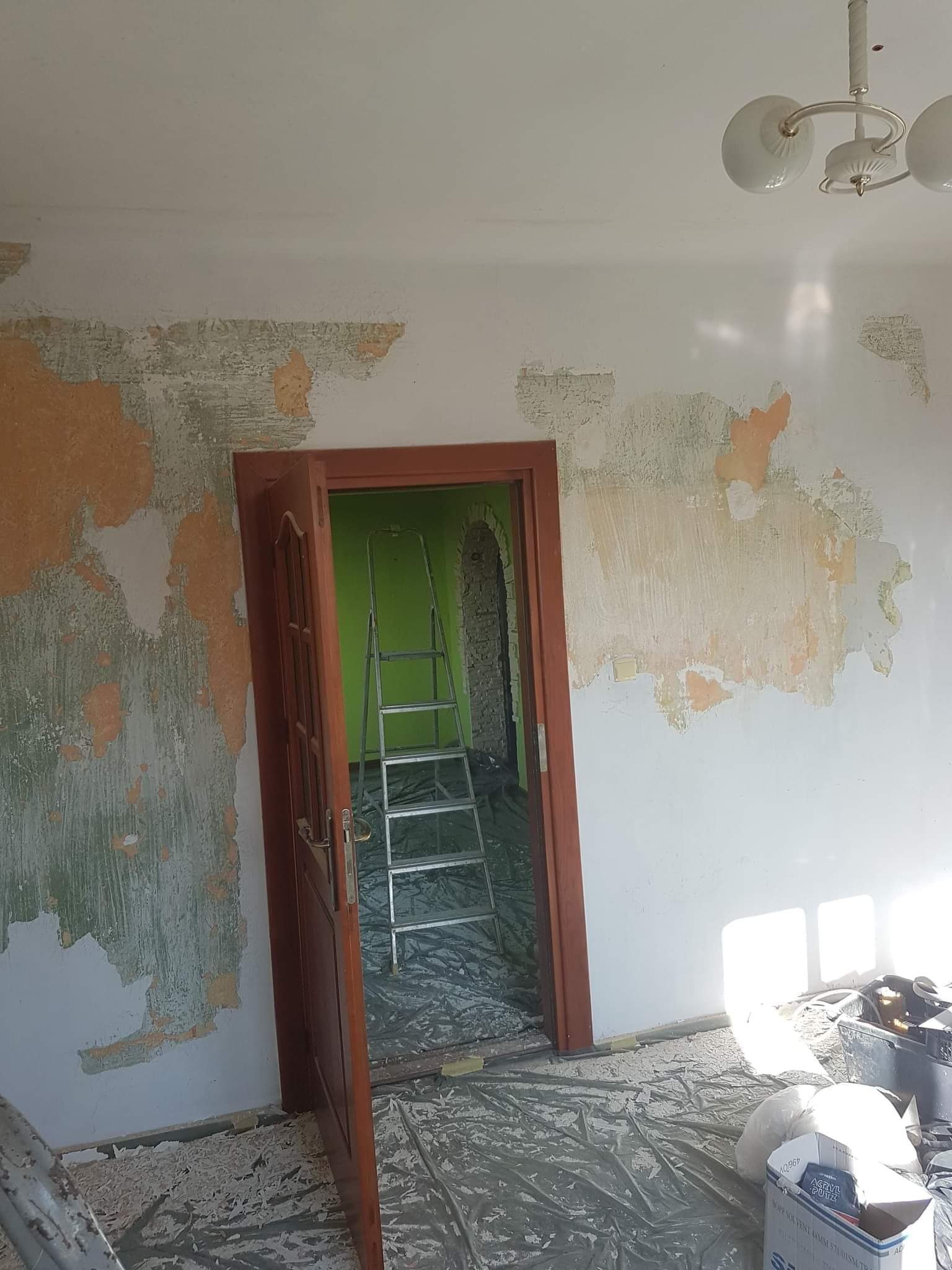 Pokój w trakcie przygotowania do malowania: ściany z resztkami starej farby i szpachli, zabezpieczona folią podłoga, aluminiowa drabina w przejściu do kolejnego pomieszczenia pomalowanego na zielono.