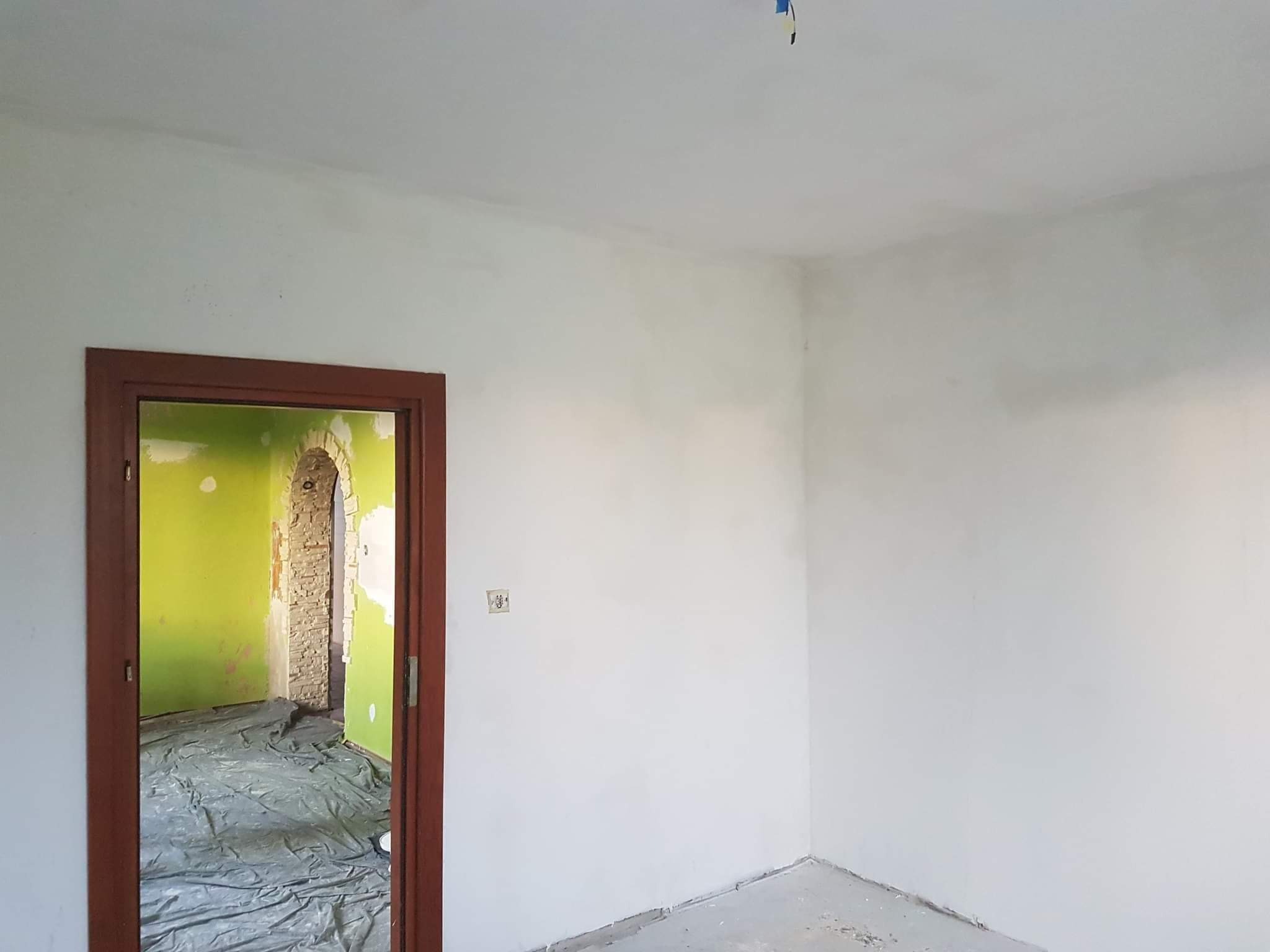 Świeżo pomalowane na biało ściany pokoju z brązową futryną drzwi, przez którą widać przygotowane do remontu wnętrze z folią ochronną na podłodze i fragmentem ściany pomalowanej na zielono.