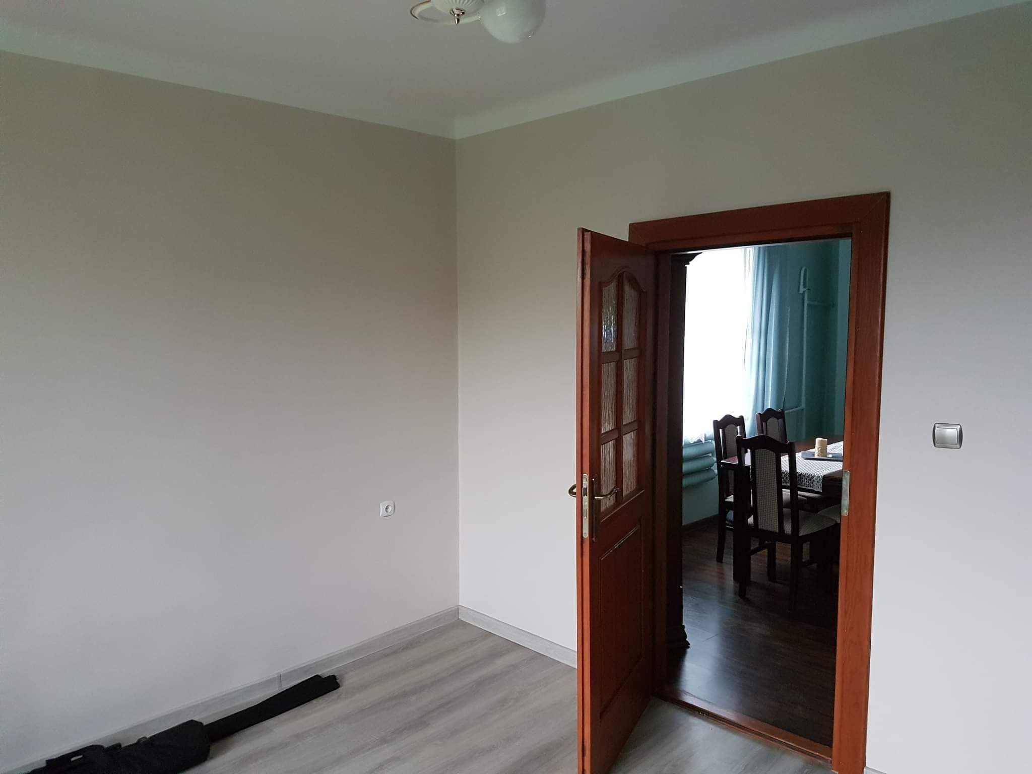 Jasne wnętrze po remoncie: ściany pomalowane na beżowo, drewniane drzwi uchylone, widoczny fragment podłogi z paneli i wnętrze drugiego pokoju z zasłonami i stołem z krzesłami.