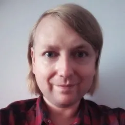 Portret uśmiechniętego mężczyzny w czerwonej koszuli w kratę na jasnoszarym tle.