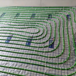 Zielone rury ogrzewania podłogowego, mocowane białymi uchwytami do folii izolacyjnej z nadrukiem siatki, przygotowane do zalania wylewką.