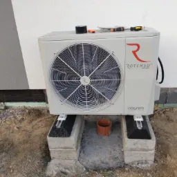 Zewnętrzna jednostka pompy ciepła marki Rotenso Aquami Series, zamontowana na betonowych bloczkach, z narzędziami (taśma, śrubokręty) na górze, widok z boku.