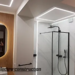 Nowoczesna łazienka z podświetlanym lustrem owalnym, kabiną prysznicową typu walk-in z czarną armaturą i liniowym oświetleniem LED w suficie i ścianach, odbicie fotografa w lustrze.