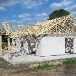 Budowa domu jednorodzinnego w stanie surowym otwartym: ściany z betonu komórkowego, drewniana konstrukcja dachu, widoczny betonowy fundament.