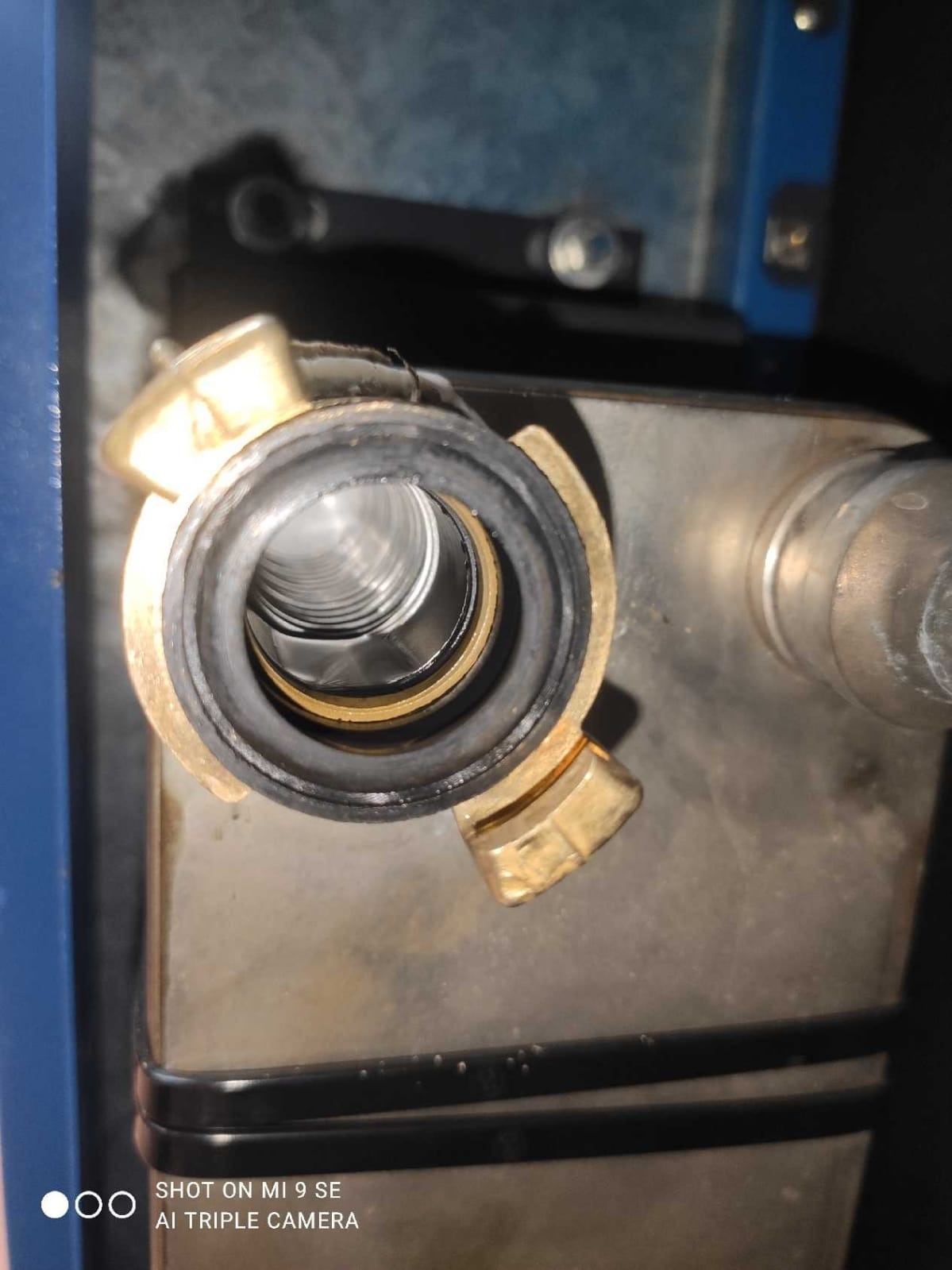 Złącze hydrauliczne z gwintem wewnętrznym, uszczelką i elementami mosiężnymi, widoczne na tle metalowej powierzchni urządzenia.