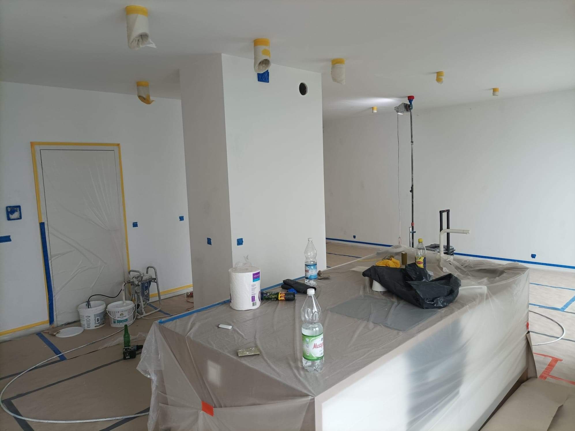 Przygotowania do malowania mieszkania: pomieszczenie zabezpieczone folią i taśmą malarską, widoczny agregat malarski, zakryta wyspa kuchenna, zabezpieczone lampy na suficie.