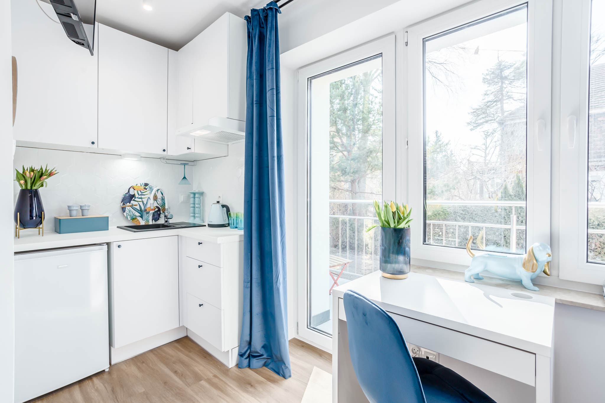 Biała, nowoczesna kuchnia z blatem roboczym pod oknem, błękitne zasłony, dekoracje w postaci wazonu z tulipanami i figurki psa.