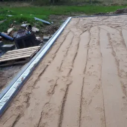 Przygotowany teren pod budowę tarasu: wyrównany piasek z widocznymi śladami, obrzeża z odpływem liniowym, w tle trawa i drzewa.