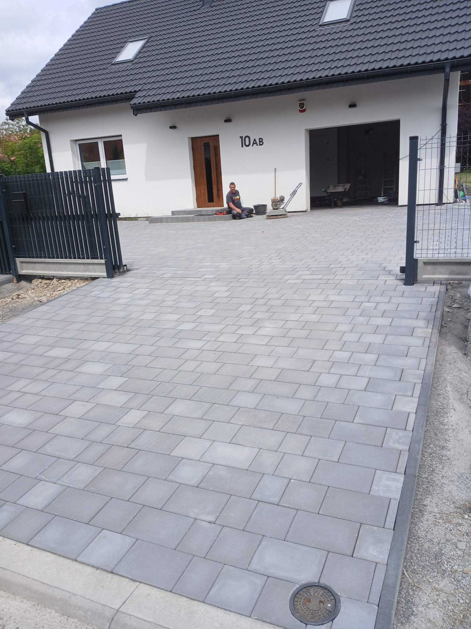 Ułożona szara kostka brukowa na podjeździe przed domem z numerem 10AB. Widoczny pracownik, ubijarka i brama wjazdowa. Efekt po wykonanej usłudze.
