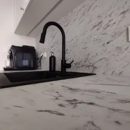 Czarny, minimalistyczny zlew i bateria na blacie z marmurowym wzorem, z widocznym dozownikiem mydła i ekspresem do kawy w tle.