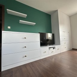 Meble Bruno spółka z o.o. - Biała zabudowa meblowa z szufladami i szafą, uzupełniona o półki wiszące na zielonej ścianie, z akcentem złotych gałek na frontach szuflad i szafek.
