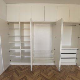 Meble Bruno spółka z o.o. - Biała szafa wnękowa z otwartymi drzwiami, ukazująca półki, kosze druciane i szuflady, na tle parkietu w jodełkę.