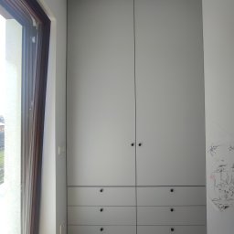 Meble Bruno spółka z o.o. - Szara szafa wnękowa z ciemnymi okrągłymi uchwytami, sześć szuflad u dołu, obok okna z widokiem na zewnątrz, na ścianie dziecięce rysunki.