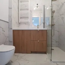 Nowoczesna łazienka z białymi marmurowymi płytkami, drewnianą szafką pod umywalką z okrągłymi złotymi uchwytami, owalnym lustrem, białą toaletą i kabiną prysznicową ze złotą armaturą.