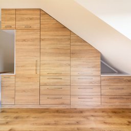 Meble Bruno spółka z o.o. - Drewniana szafa wnękowa na poddaszu we Wrocławiu, dopasowana do skosu dachu, zintegrowana z ramą łóżka, widoczna na tle ściany z tapetą.