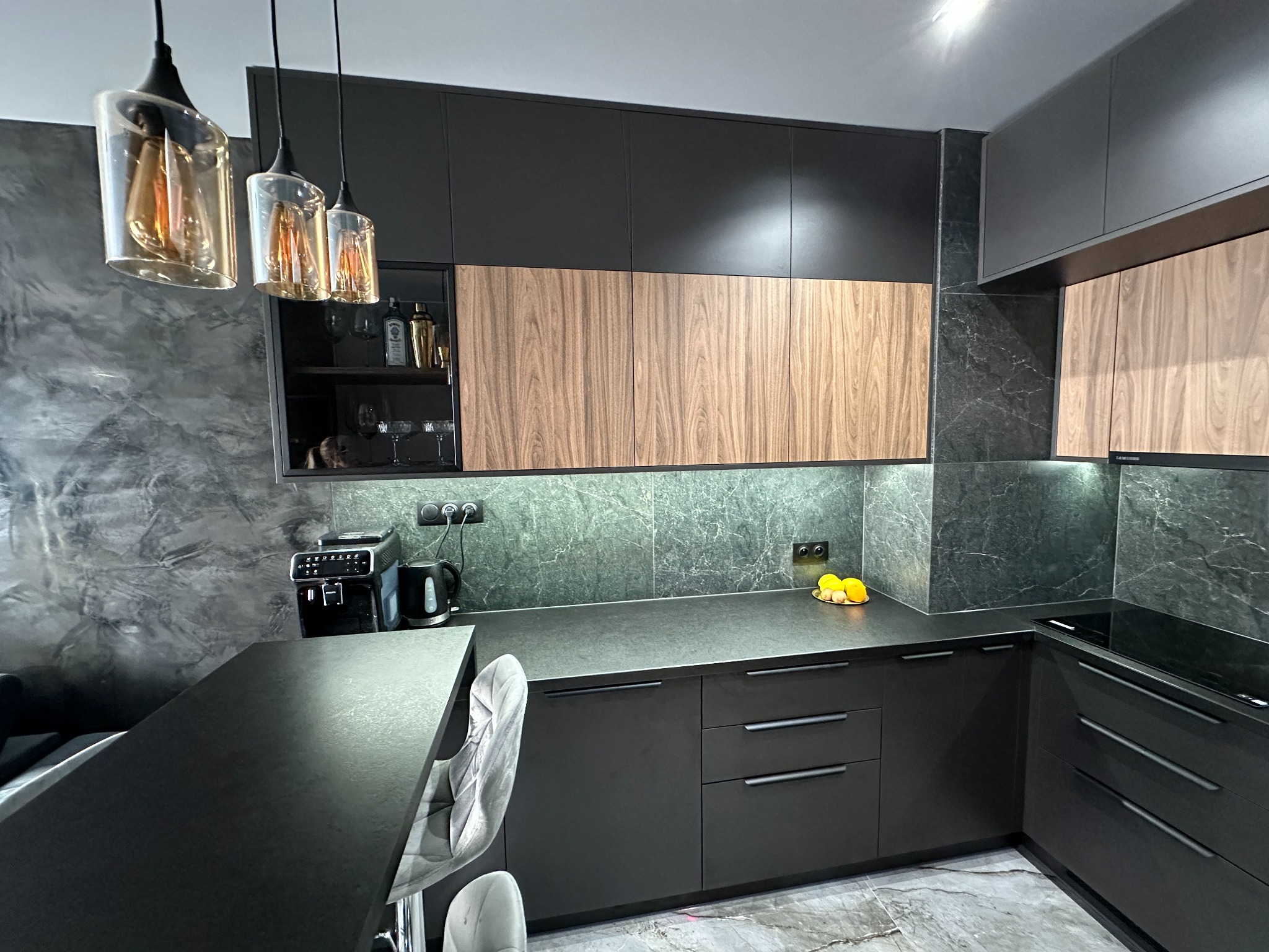 Nowoczesna kuchnia z czarnymi matowymi szafkami dolnymi i górnymi, drewnianymi frontami szafek wiszących, marmurowym blatem i ścianą, oraz trzema wiszącymi lampami z bursztynowego szkła.