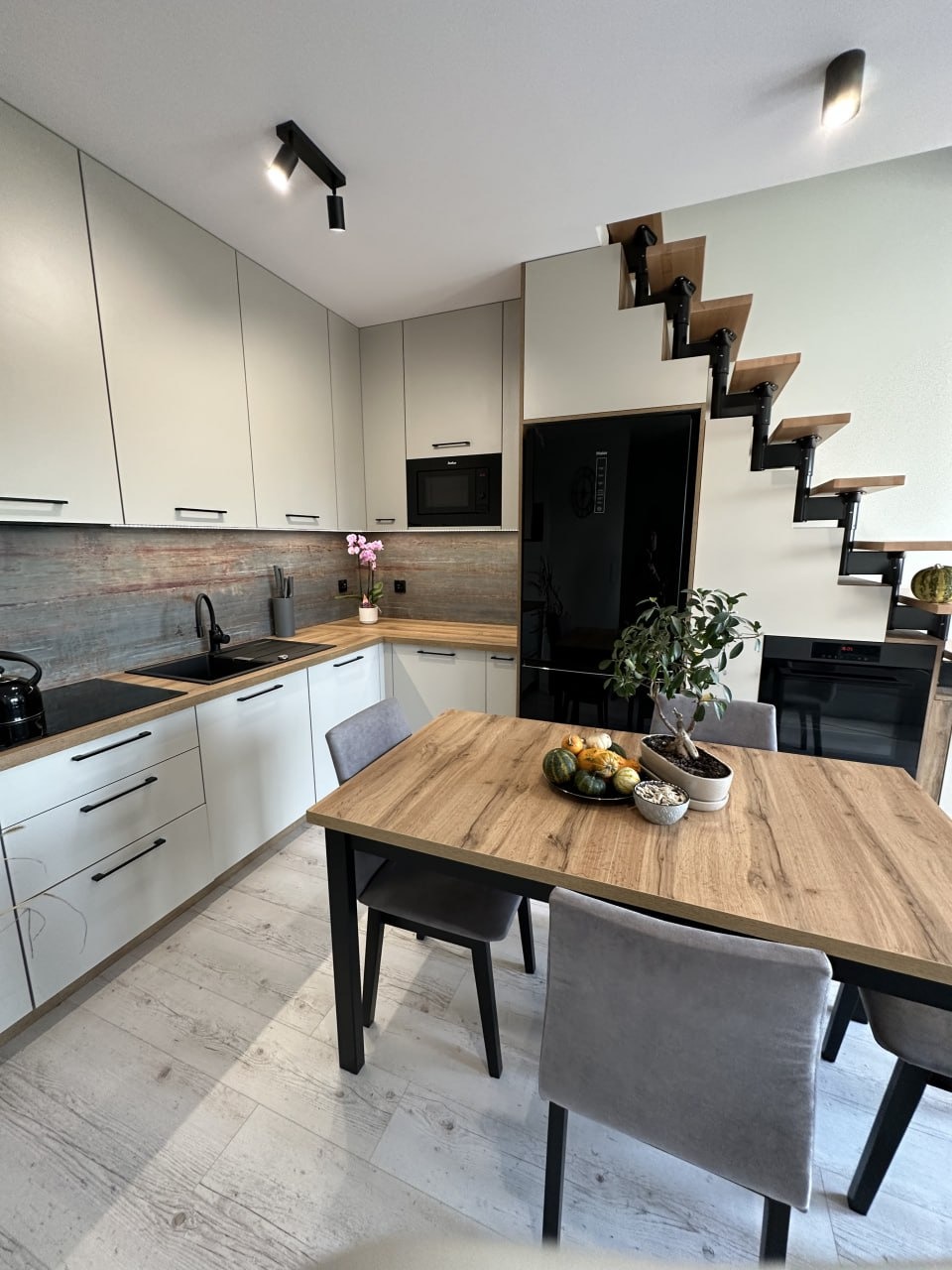 Jasna kuchnia z drewnianym blatem, czarnymi akcentami i schodami prowadzącymi na górę, stół z dekoracyjnymi dyniami i bonsai.