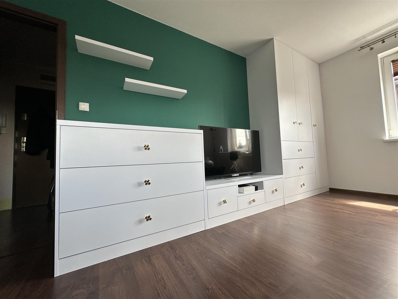 Biała zabudowa meblowa z szufladami i szafą, uzupełniona o półki wiszące na zielonej ścianie, z akcentem złotych gałek na frontach szuflad i szafek.