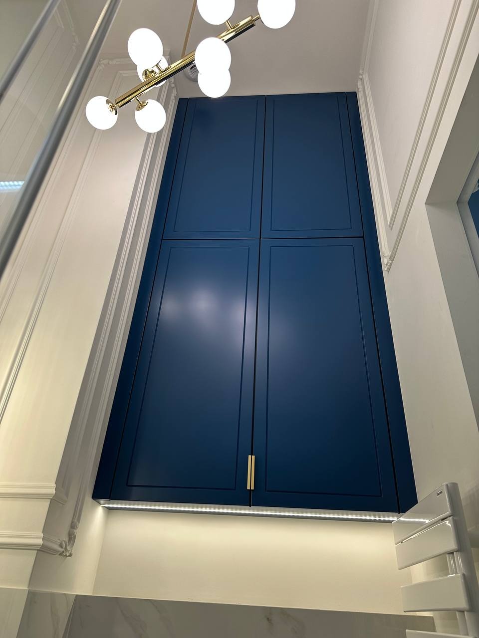 Wysoka, zabudowana szafa wnękowa w kolorze granatowym z złotymi uchwytami, oświetlona od dołu, w nowoczesnej łazience z dekoracyjną sztukaterią.