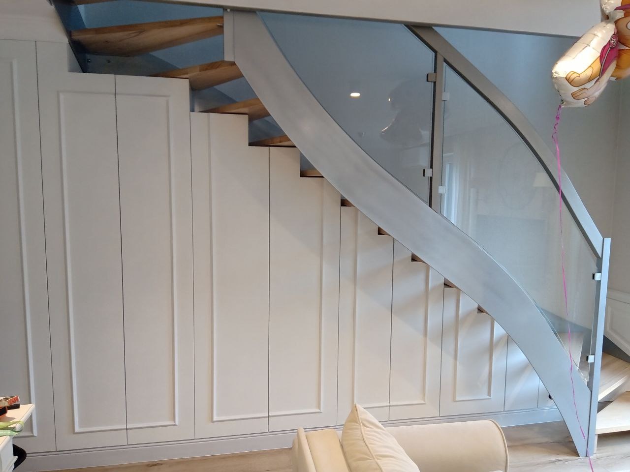 Białe szafy wnękowe z frezowanymi frontami pod schodami z drewnianymi stopniami i szklaną balustradą z metalowymi elementami.