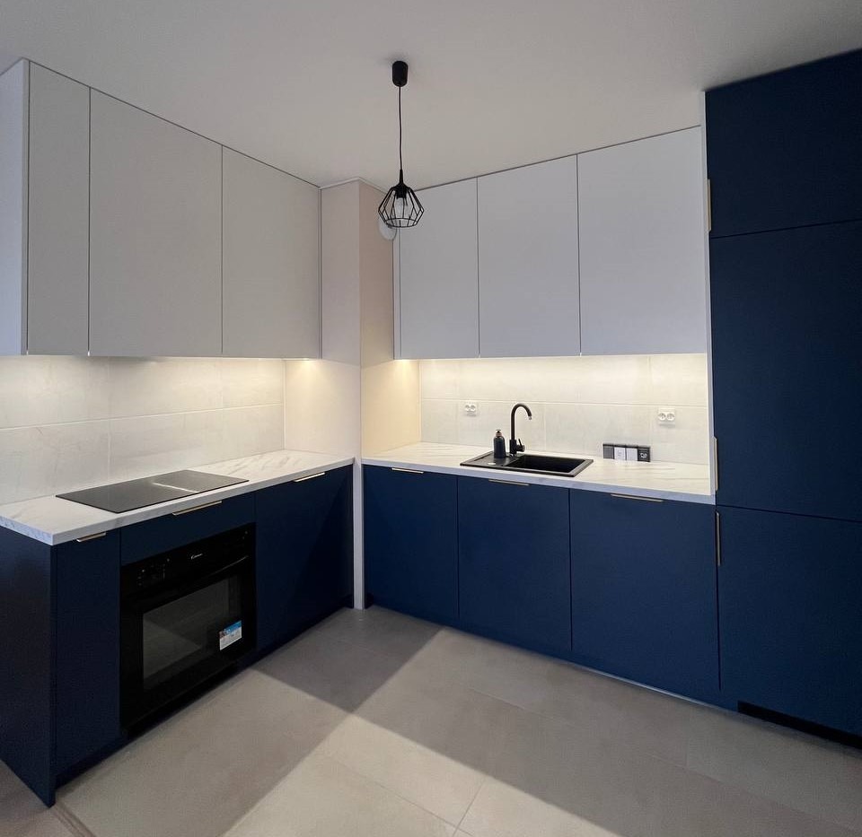 Nowoczesna kuchnia z białymi górnymi szafkami i granatowymi dolnymi, marmurowym blatem, czarnym zlewem i płytą indukcyjną, oświetleniem LED pod szafkami i geometryczną lampą wiszącą.